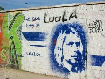 A blue stencil portrait of Kurt Cobain on a white wall covered in graffiti, including the name "Lucila", dates, and surrounding tags.