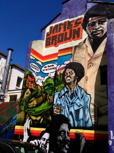 A tall James Brown mural on the side of a building, with large red "JAMES BROWN" lettering, portraits, comic-style figures, and a speech bubble reading "That's because he's the Godfather."