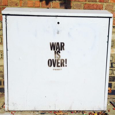 The words "War is over! If you want it." stencilled on a plain white street cabinet against a brick wall.