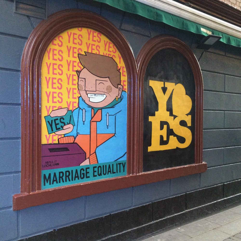 A pub window painted with a comic-style figure posting a "YES" ballot into a box against a background of repeated "YES" text and the words "MARRIAGE EQUALITY", beside a second window reading "YES"; signed "Niall O'Lochlainn".