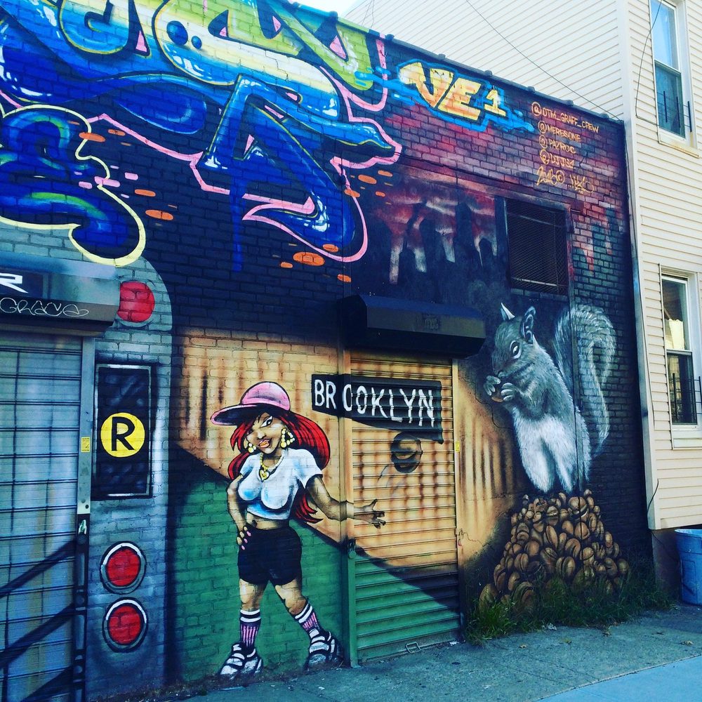 A stylised woman in a pink cap and a grey squirrel beside the word "BROOKLYN", painted with colourful graffiti on a wall beside a shuttered doorway.