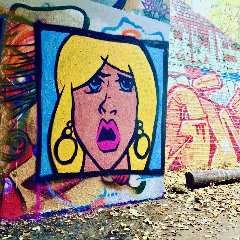 A Lichtenstein-style blonde face with wide eyes and an open pink mouth, framed in a square panel on a graffiti-covered wall.