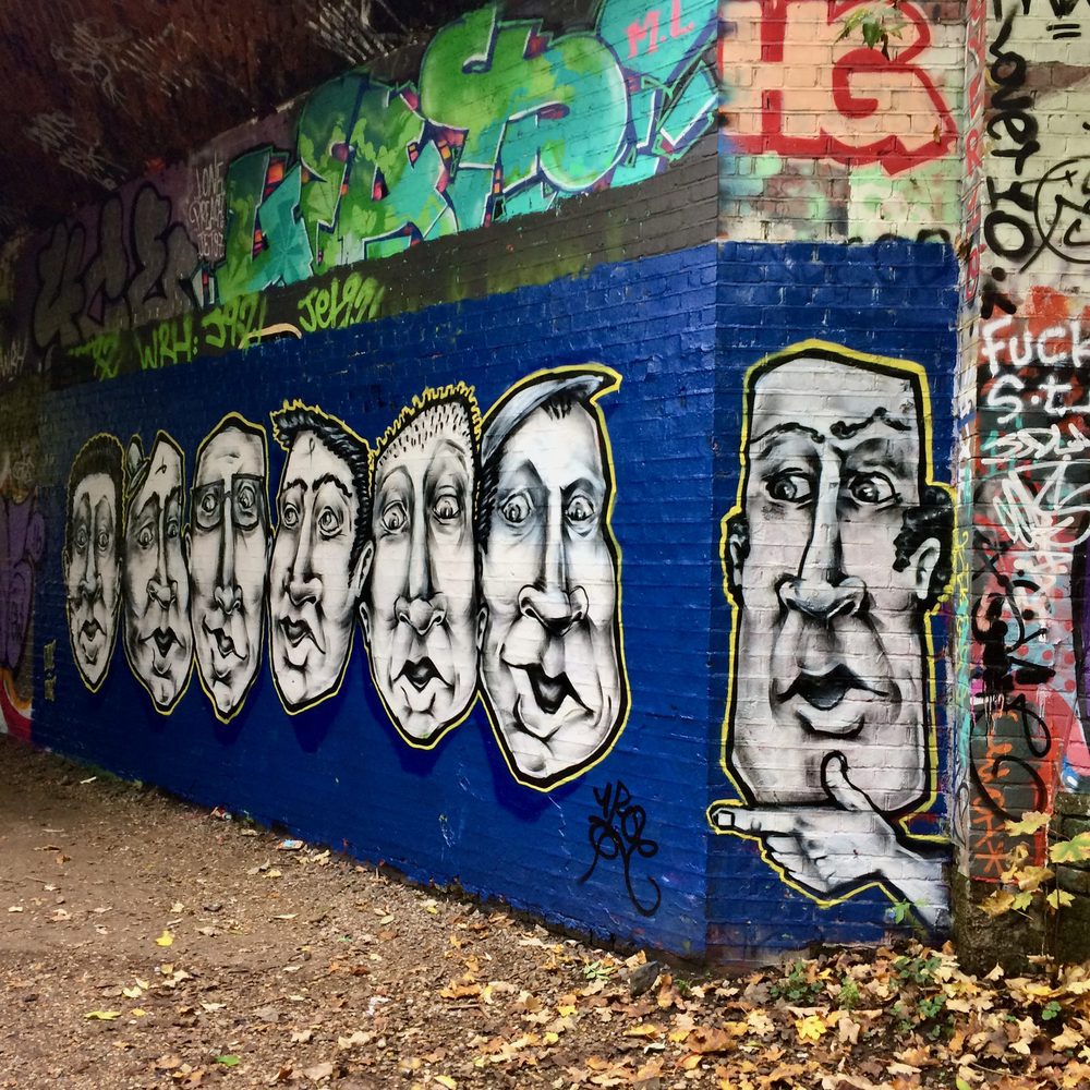 A row of monochrome stylised faces painted on a blue brick wall beneath a bridge, surrounded by graffiti.