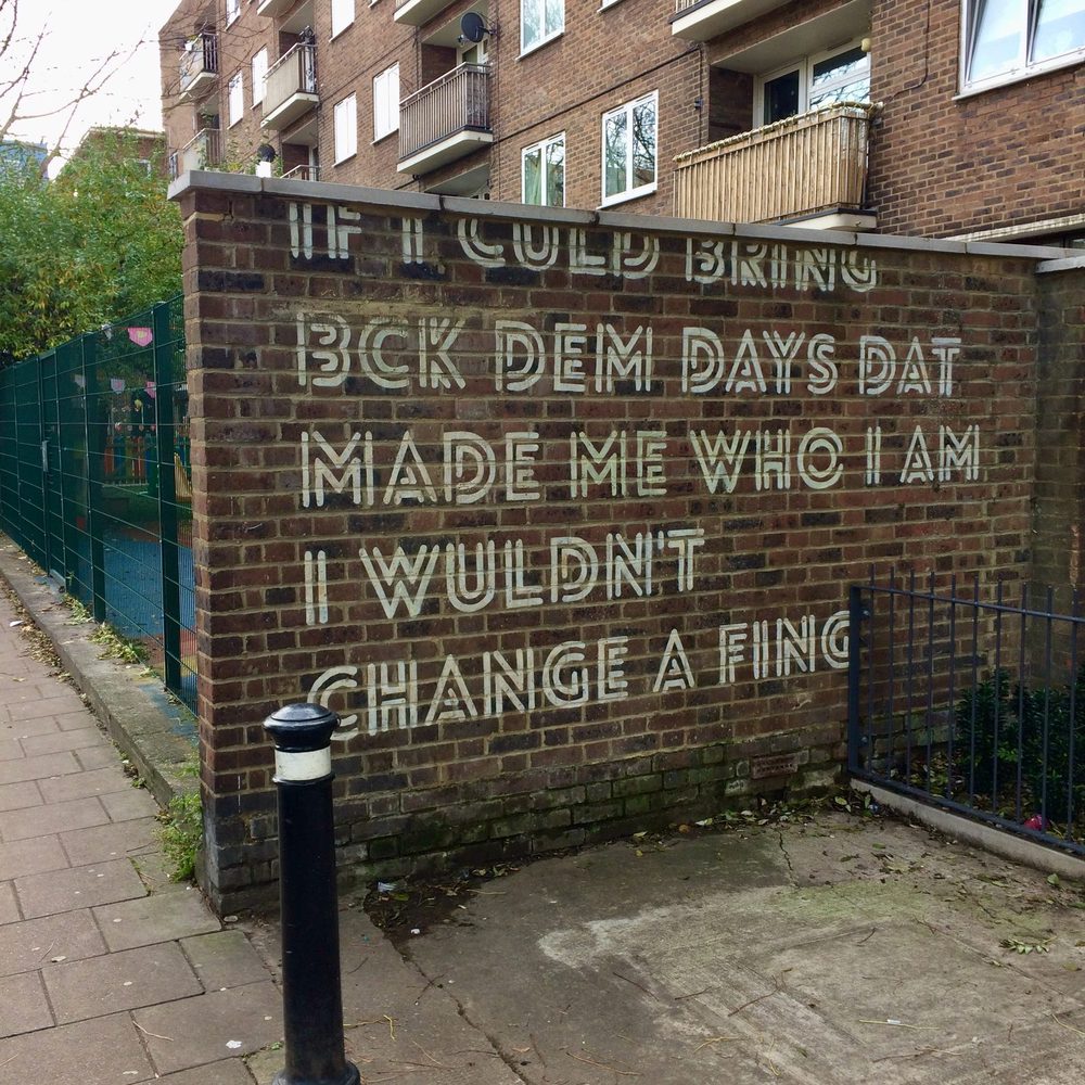 Large white text reading "If I could bring back dem days dat made me who I am I wouldn't change a fing." painted on a brick wall.