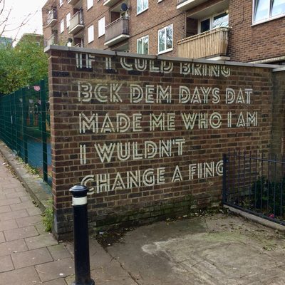 Large white text reading "If I could bring back dem days dat made me who I am I wouldn't change a fing." painted on a brick wall.