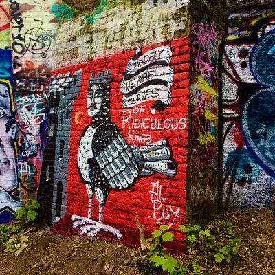 A black-and-white bird holding a scroll that reads "Today we are slaves of Ridiculous Kings", painted on a red brick wall.