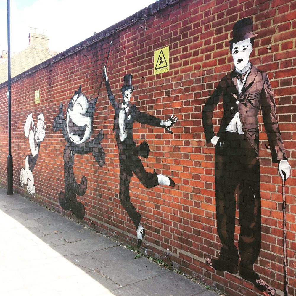 A large Charlie Chaplin figure holding a cane, alongside Felix the Cat and other silent-era comic figures, painted on a red brick wall.