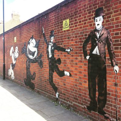 A large Charlie Chaplin figure holding a cane, alongside Felix the Cat and other silent-era comic figures, painted on a red brick wall.