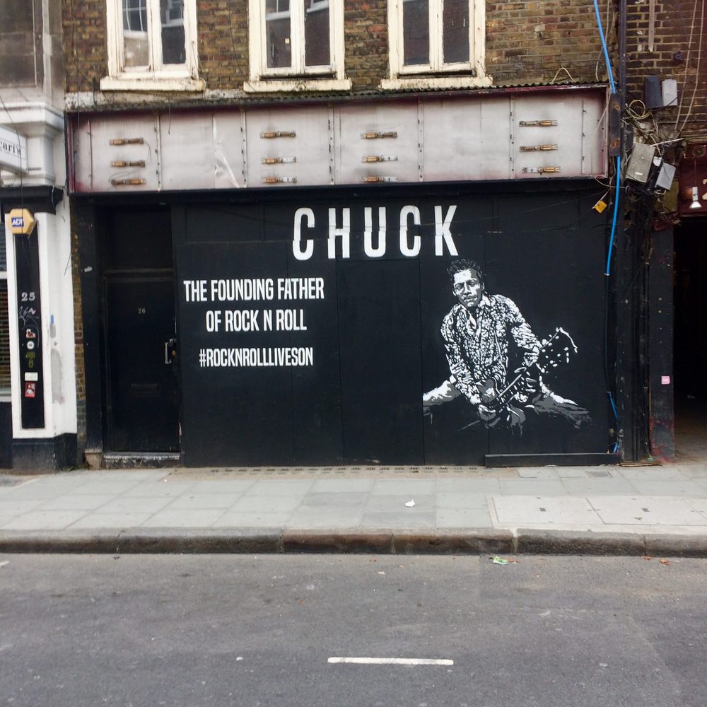 A white stencil portrait of Chuck Berry playing guitar, beside text reading "CHUCK / THE FOUNDING FATHER OF ROCK N ROLL" and the hashtag "#ROCKNROLLLIVESON", on a black storefront.