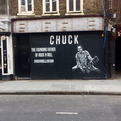 A white stencil portrait of Chuck Berry playing guitar, beside text reading "CHUCK / THE FOUNDING FATHER OF ROCK N ROLL" and the hashtag "#ROCKNROLLLIVESON", on a black storefront.