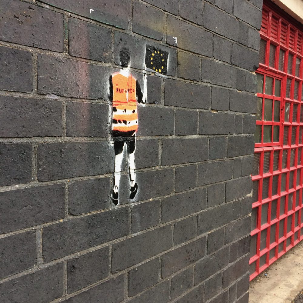 A small stencil of a figure in an orange high-visibility vest reading "FUTURE", with a partial EU flag above, on a dark brick wall beside a red metal window grille.