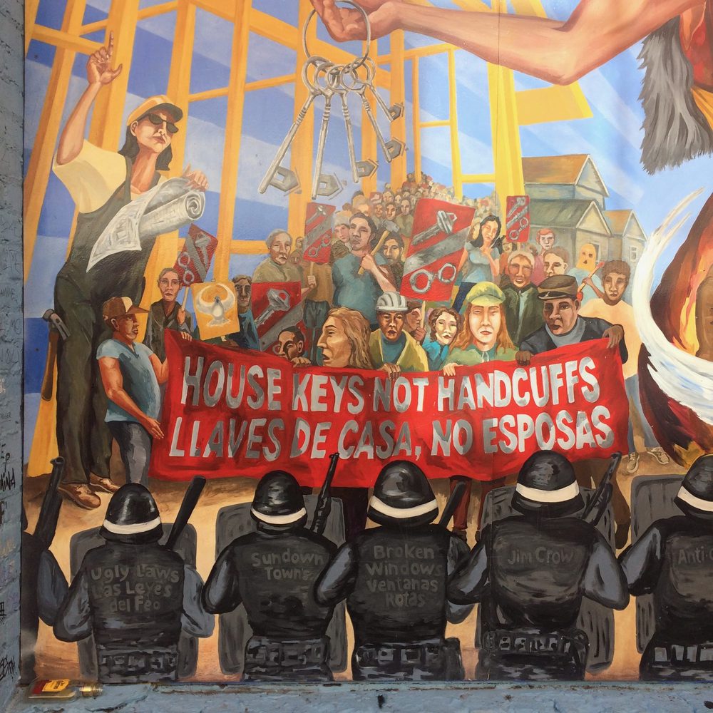 A crowd holding a red banner reading "House keys not handcuffs / Llaves de casa, no esposas", painted as a protest mural.