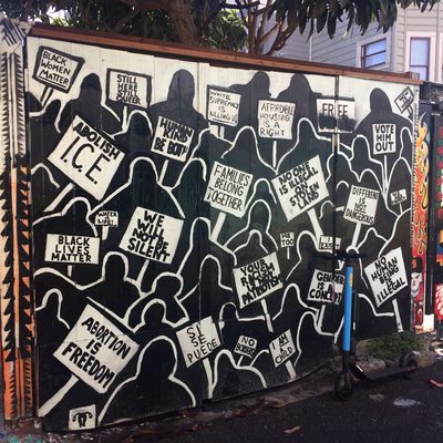 Silhouetted people holding protest signs including "Abolish ICE", "Black Lives Matter", "Families Belong Together", "Abortion is Freedom", and "No One is Illegal", painted as a black-and-white mural.