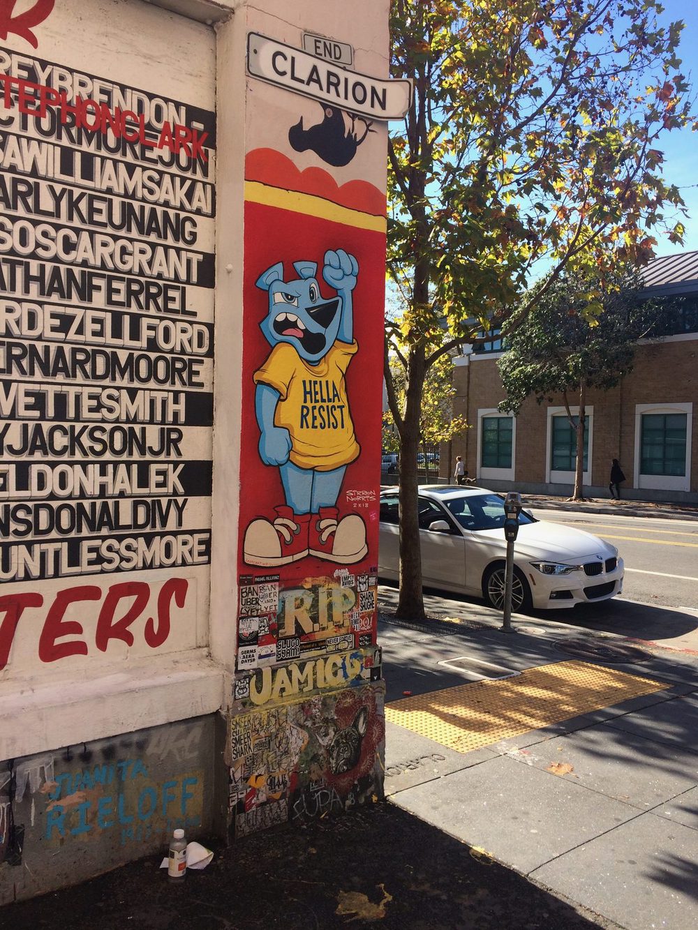 A blue stylised figure in a yellow "Hella Resist" shirt, painted on a corner beside a Clarion street sign.