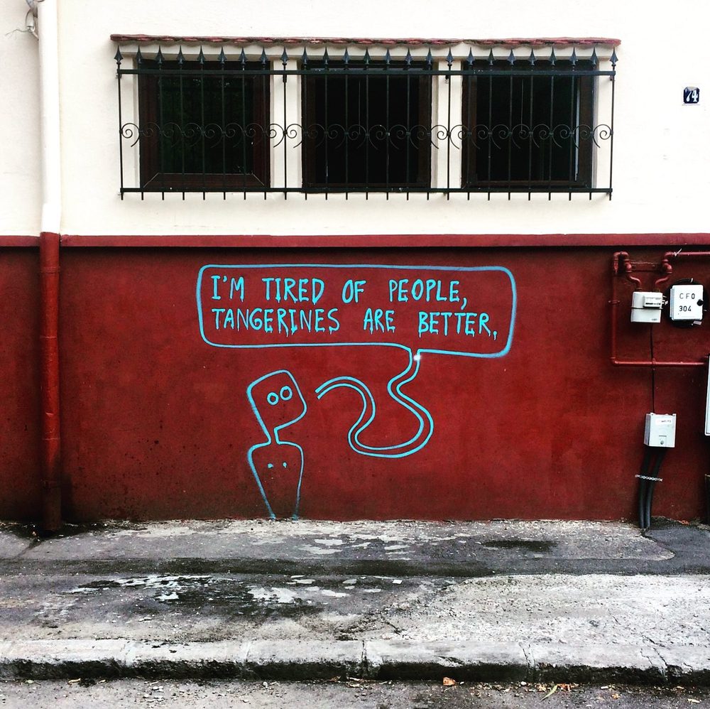 A simple blue figure saying "I'm tired of people, tangerines are better." painted on a red lower wall beneath barred windows.