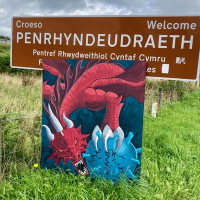 A painted red dragon fighting a blue spiky coronavirus-like creature, on a freestanding panel in front of the Welsh bilingual welcome sign for Penrhyndeudraeth, in long grass.