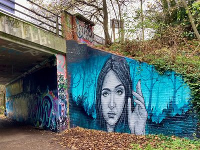 A large realistic woman's face and raised finger against a blue background, painted under a bridge beside surrounding graffiti.