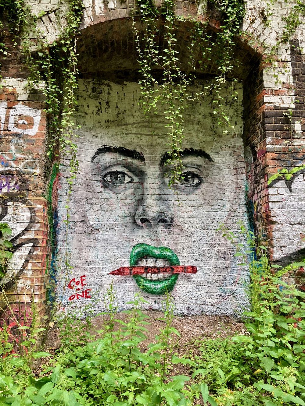 A face with green lips holding a red pencil between its teeth, painted on a brick archway and partly covered by ivy.
