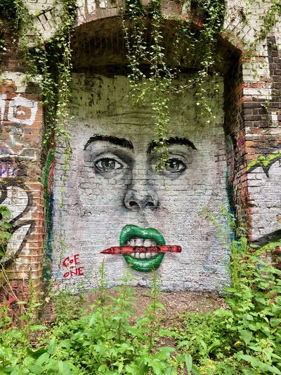 A face with green lips holding a red pencil between its teeth, painted on a brick archway and partly covered by ivy.