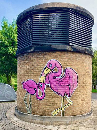 Two pink flamingos painted across the curved brick face of a round ventilation structure, beneath black louvered vents.