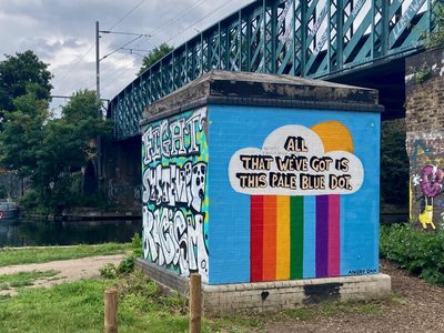 Rainbow stripes and the words "All that we've got is this pale blue dot." painted on a blue canal-side utility box.