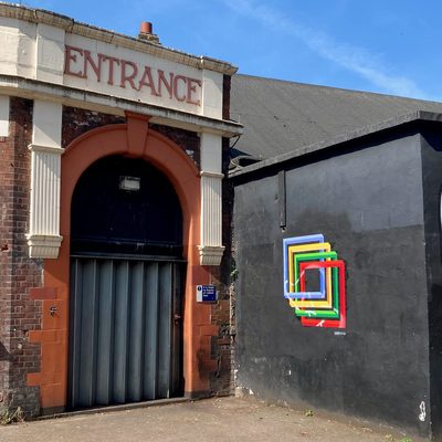 Stacked, brightly coloured square frames painted on a dark wall beside an old entrance archway.