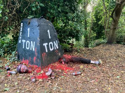 A woodland installation with a black one-ton weight, red paint, mannequin body parts, and spray cans on the ground.