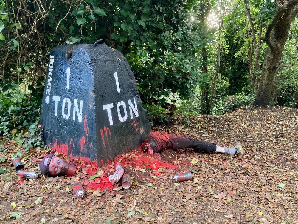 A woodland installation with a black one-ton weight, red paint, mannequin body parts, and spray cans on the ground.