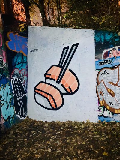 Two salmon nigiri sushi pieces held by black chopsticks, painted on a white wall and photographed at night.
