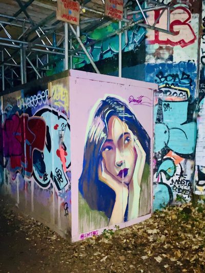 A woman resting her face on her hand, painted on a pink fence in a graffiti-covered underpass.