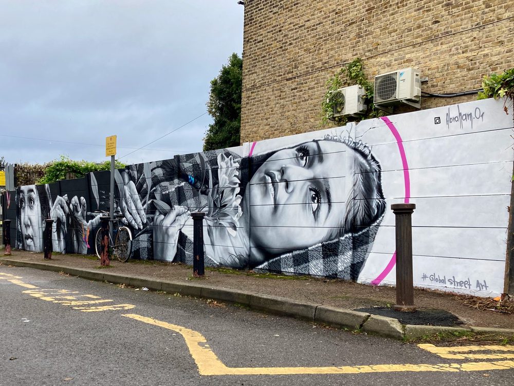 A person looking upward, surrounded by hands, flowers, and pink accent arcs, painted in black and white as a long roadside mural; signed "@abraham.or".