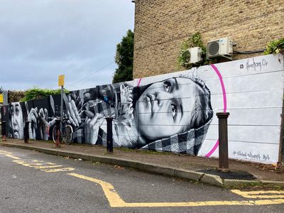 A person looking upward, surrounded by hands, flowers, and pink accent arcs, painted in black and white as a long roadside mural; signed "@abraham.or".