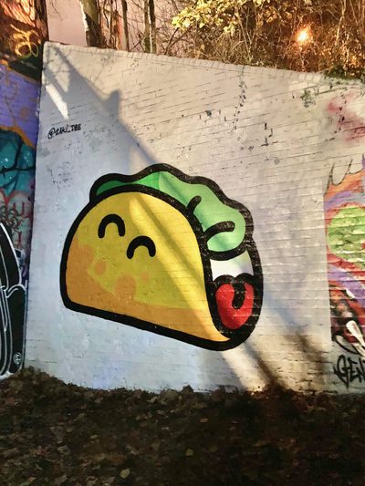 A stylised taco with green filling and red sauce, painted on a white wall and photographed at night.