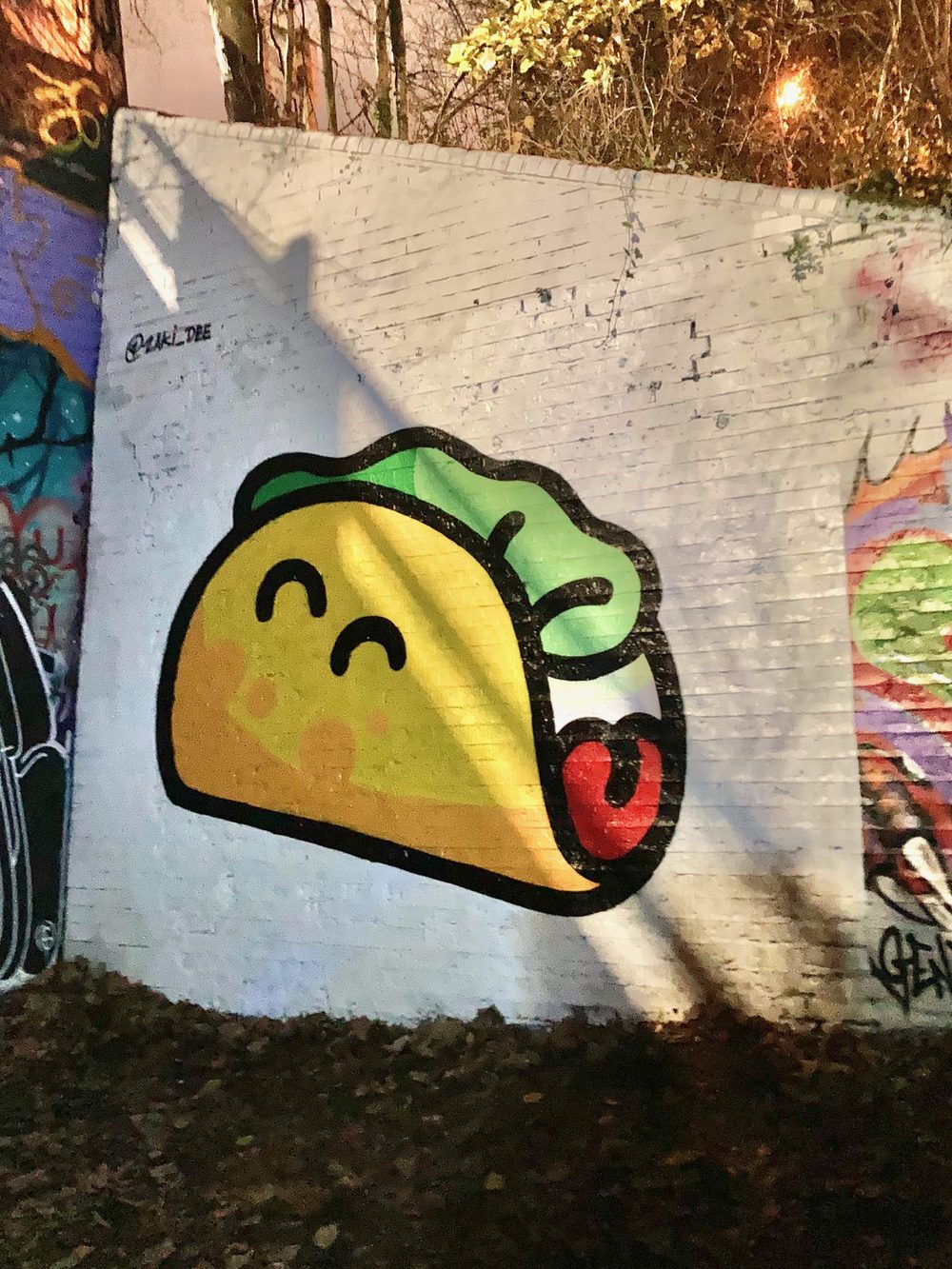 A stylised taco with green filling and red sauce, painted on a white wall and photographed at night.