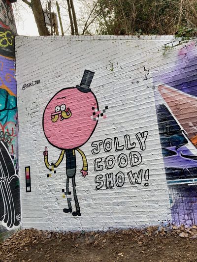 A pink round-headed stylised figure in a top hat beside the words "Jolly good show!" painted on a white brick wall; signed "@zal.188".