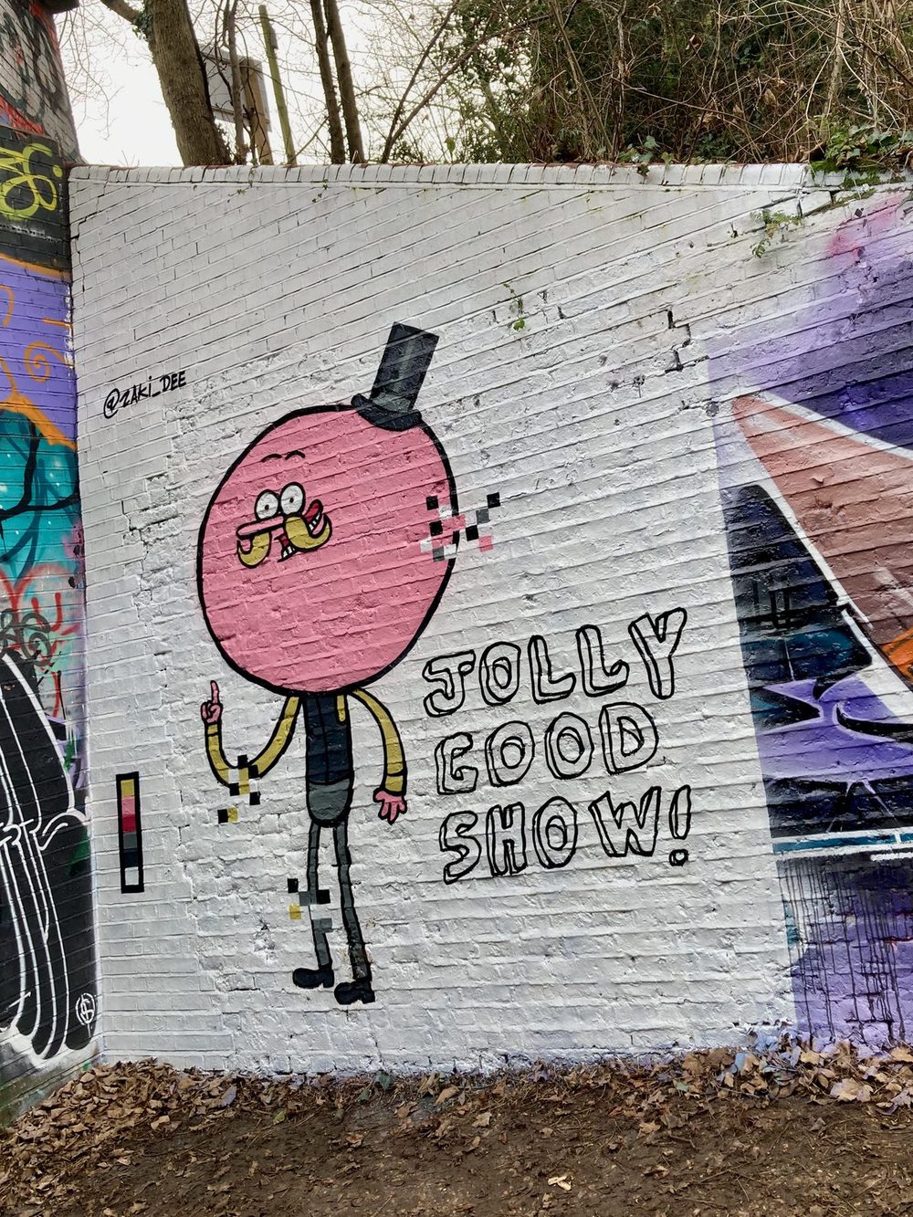 A pink round-headed stylised figure in a top hat beside the words "Jolly good show!" painted on a white brick wall; signed "@zal.188".