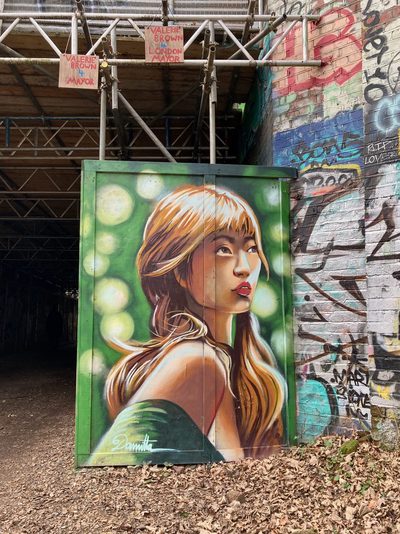 A painted portrait of a young woman with long blonde hair against a green background of bokeh-like circles, on a green doorway beneath a bridge; small "Valerie Brown for Mayor" campaign signs hang from the structure above.