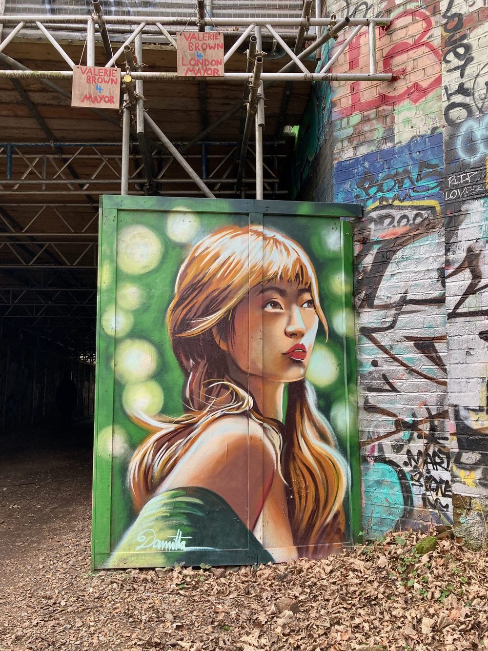 A painted portrait of a young woman with long blonde hair against a green background of bokeh-like circles, on a green doorway beneath a bridge; small "Valerie Brown for Mayor" campaign signs hang from the structure above.