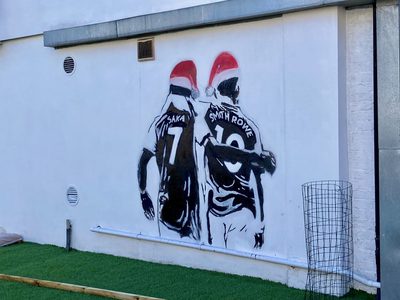 Two footballers in Santa hats with "Saka 7" and "Smith Rowe 10" on their shirts, painted on a white wall.