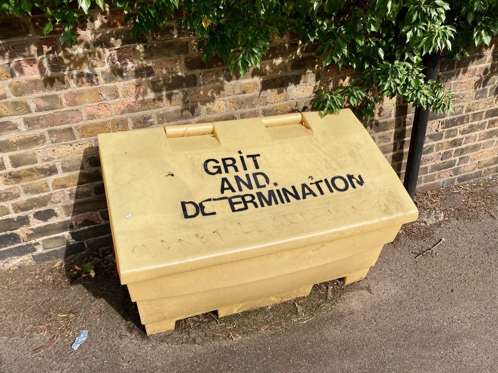 The words "Grit and determination." on a yellow grit bin beside a brick wall.