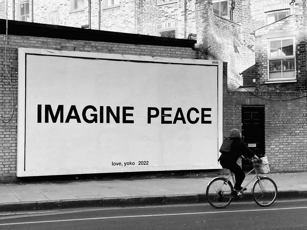 A black-and-white billboard-style artwork reading "Imagine peace" and "love, yoko 2022", with a cyclist passing by.