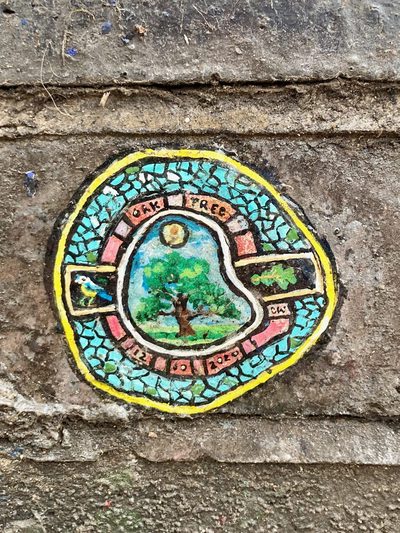 A small circular mosaic-style painting of an oak tree and bird on a stone wall, with the words "Oak Tree" around the border.