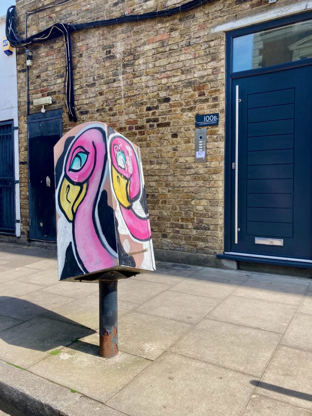 Two pink flamingo heads painted on a freestanding street box outside a brick building.