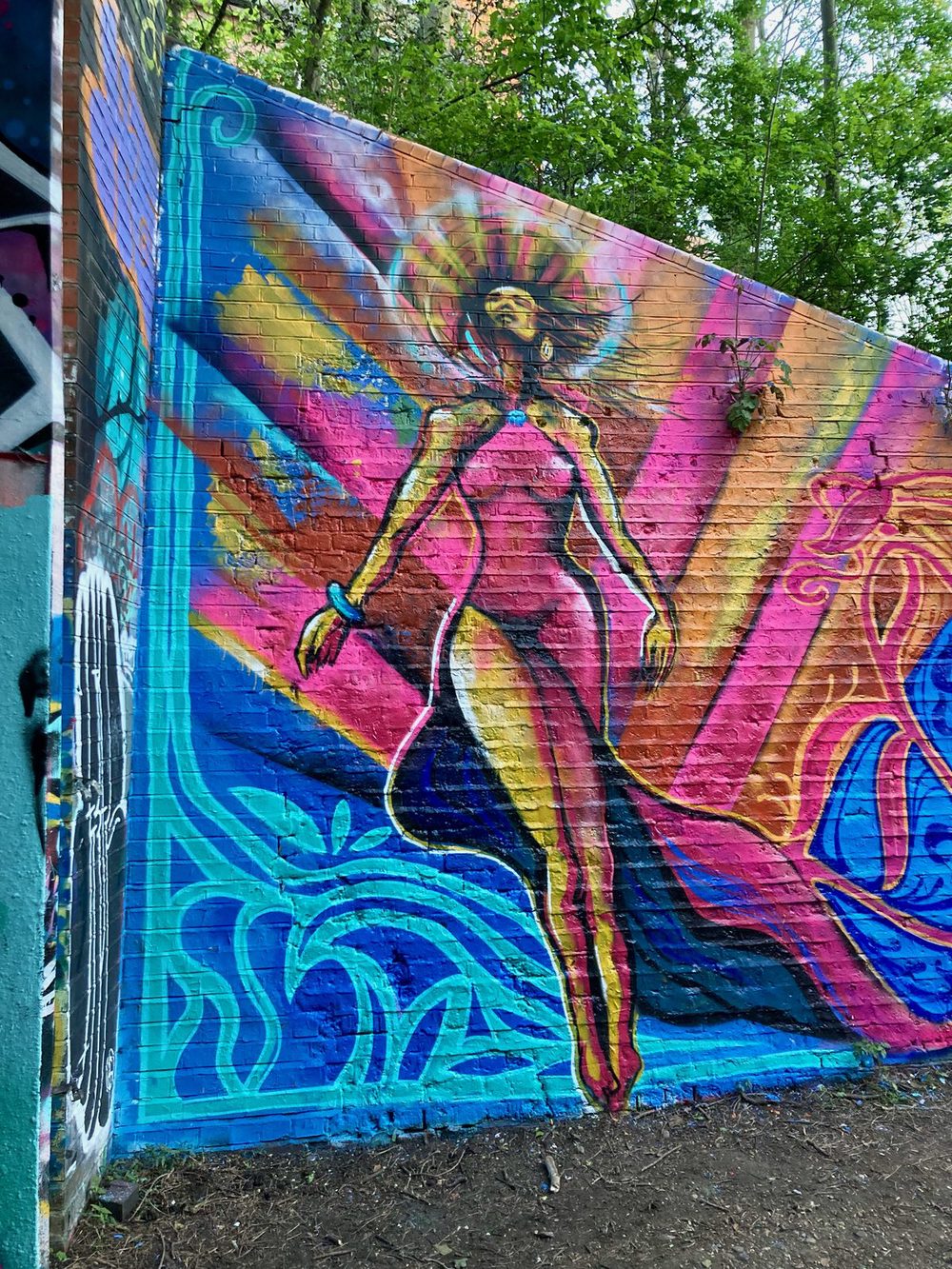 A tall stylised figure with long arms against blue, pink, and orange shapes, painted on a colourful brick wall.