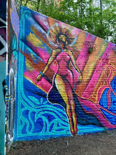 A tall stylised figure with long arms against blue, pink, and orange shapes, painted on a colourful brick wall.