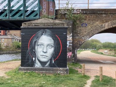 A greyscale portrait of a woman inside a red circle, painted on a black brick structure beside a canal bridge; signed "@blackmonument".