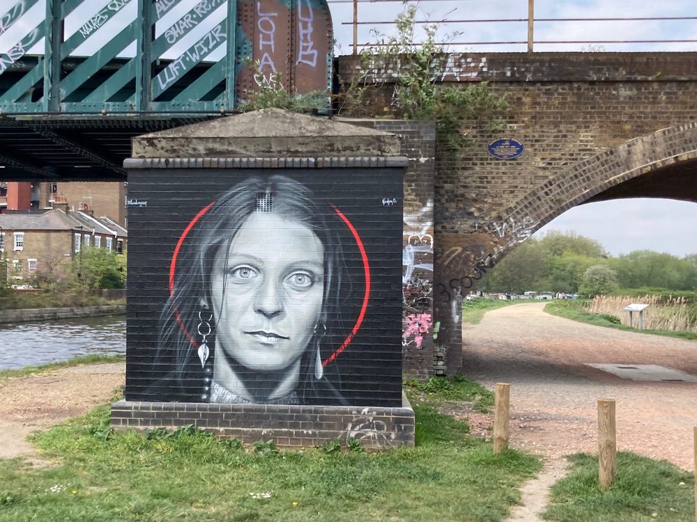 A greyscale portrait of a woman inside a red circle, painted on a black brick structure beside a canal bridge; signed "@blackmonument".