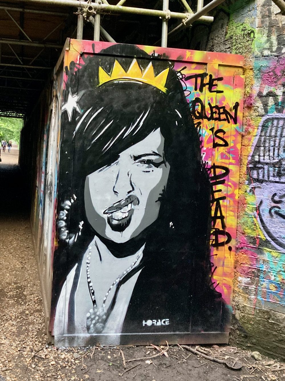 A black-and-white crowned face of Amy Winehouse beside the words "The queen is dead." painted on a street box beneath a bridge; signed "Horace".