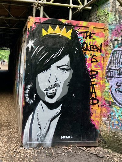 A black-and-white crowned face of Amy Winehouse beside the words "The queen is dead." painted on a street box beneath a bridge; signed "Horace".