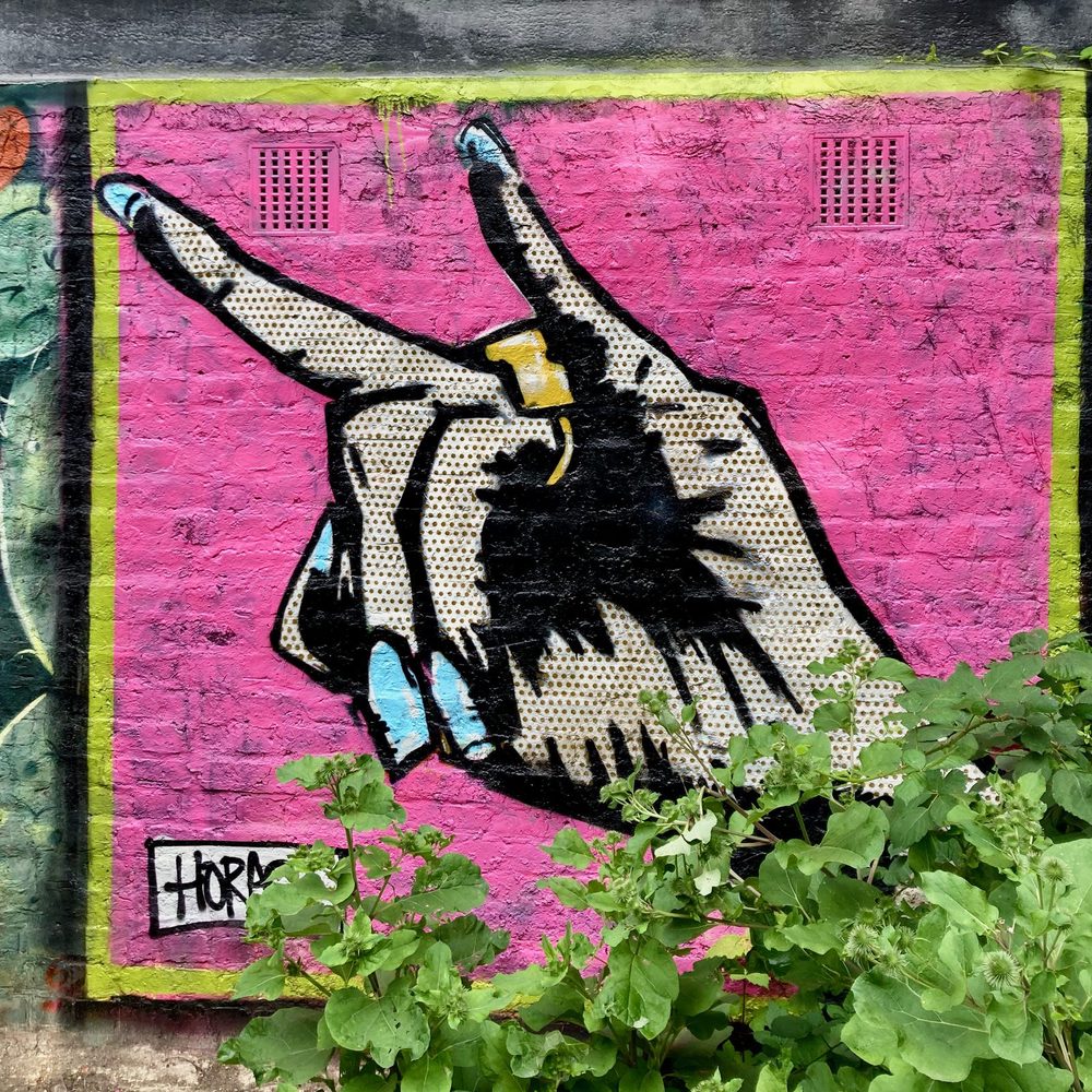 A large Lichtenstein-style hand making a devil-horns gesture, with Ben-Day dots, painted on a pink wall; signed "Horace".